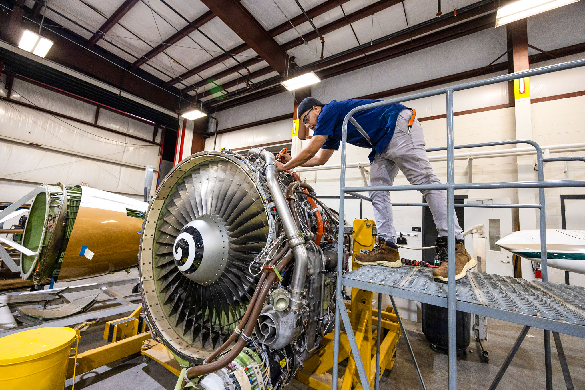 Technician standing on a metal platform inspects and works on a large exposed jet engine, reaching into its wiring and components inside an aviation training workshop.